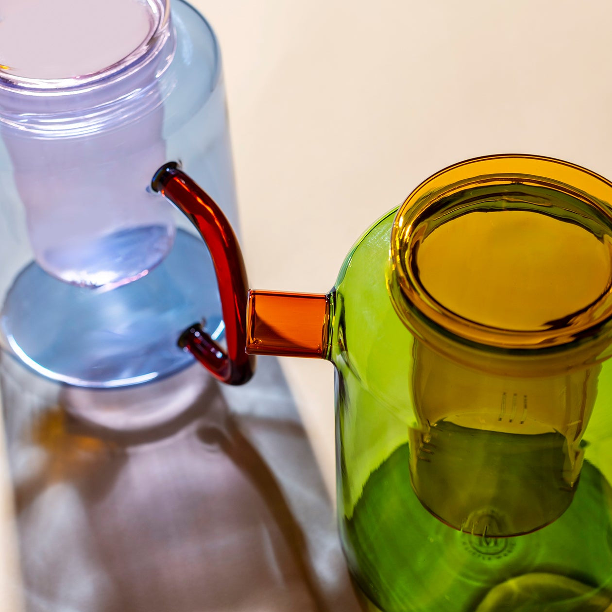 Close-up of colorful glass smoking accessories on a light beige surface, featuring a translucent green bowl with a yellow rim, a blue chamber, and a red mouthpiece.