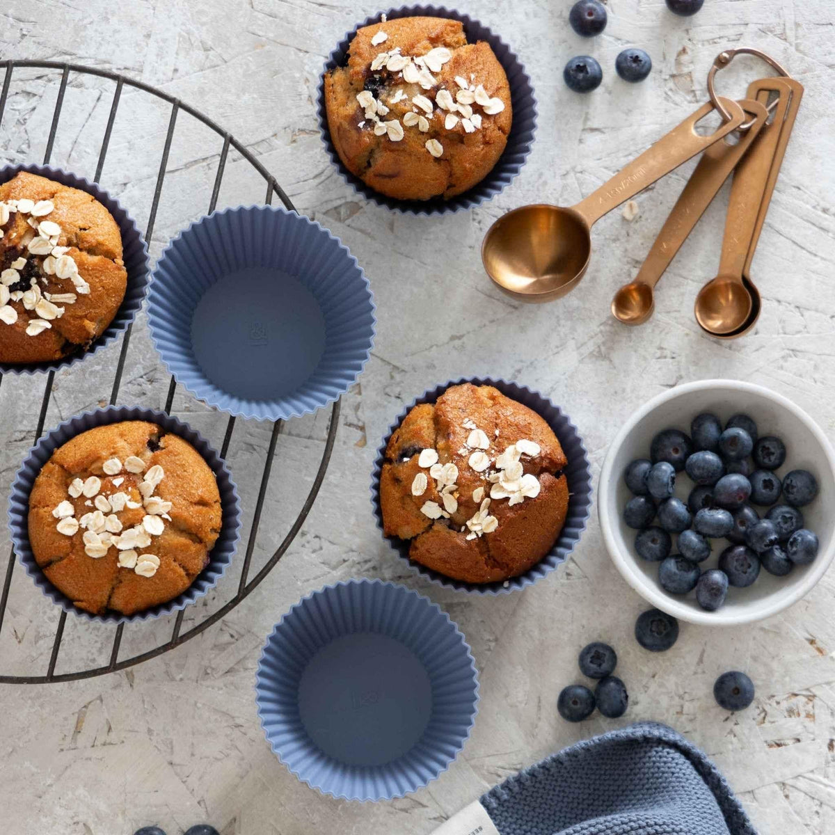 Blueberry silicone muffin cups beside oat muffins, fresh blueberries and brass measuring spoons