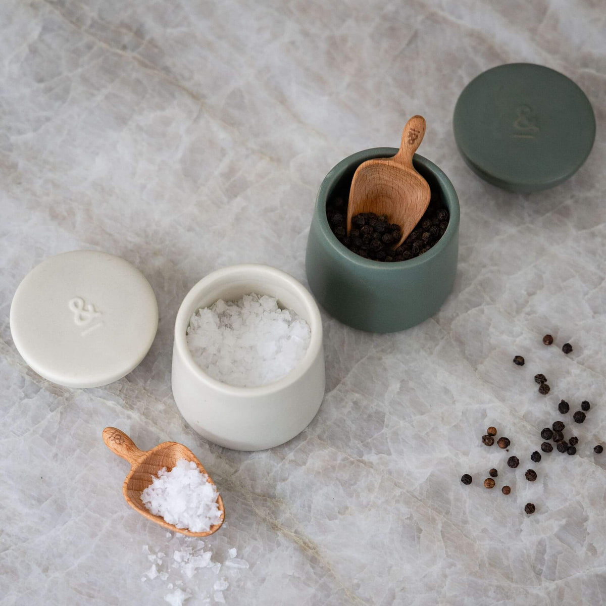Both ceramic pods open on marble, one holding flaked salt and one peppercorns, with wooden scoops