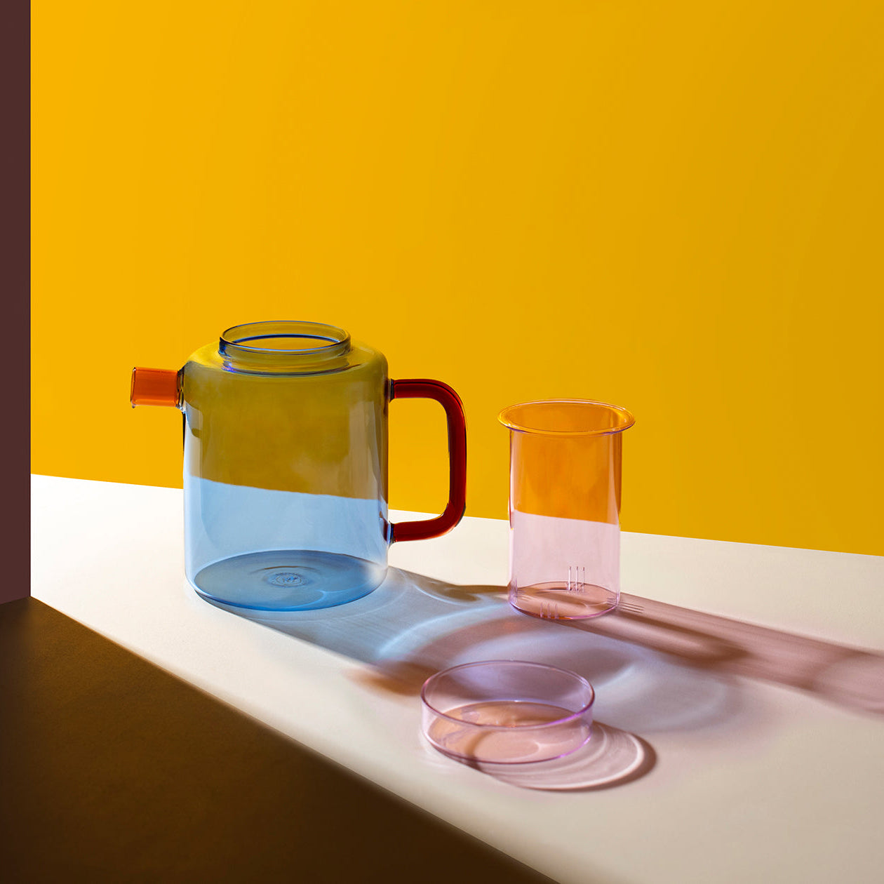 A translucent plastic teapot and matching glass cups in a minimalist studio setup with a bright yellow background and white surface, casting dramatic shadows.