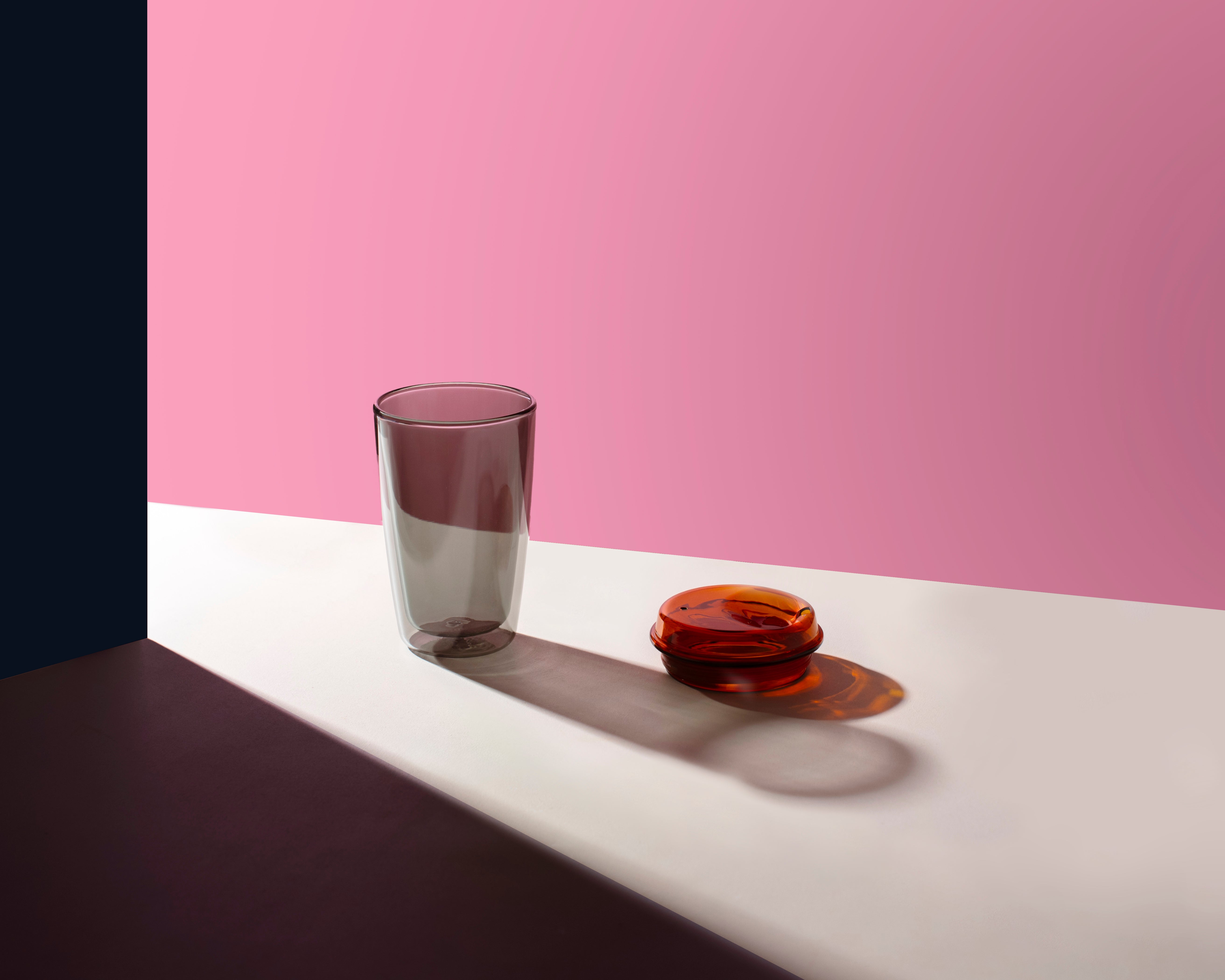 A minimalist studio still life featuring a translucent smoky gray glass tumbler and a small amber glass dish, set on a white surface against a solid pink background.