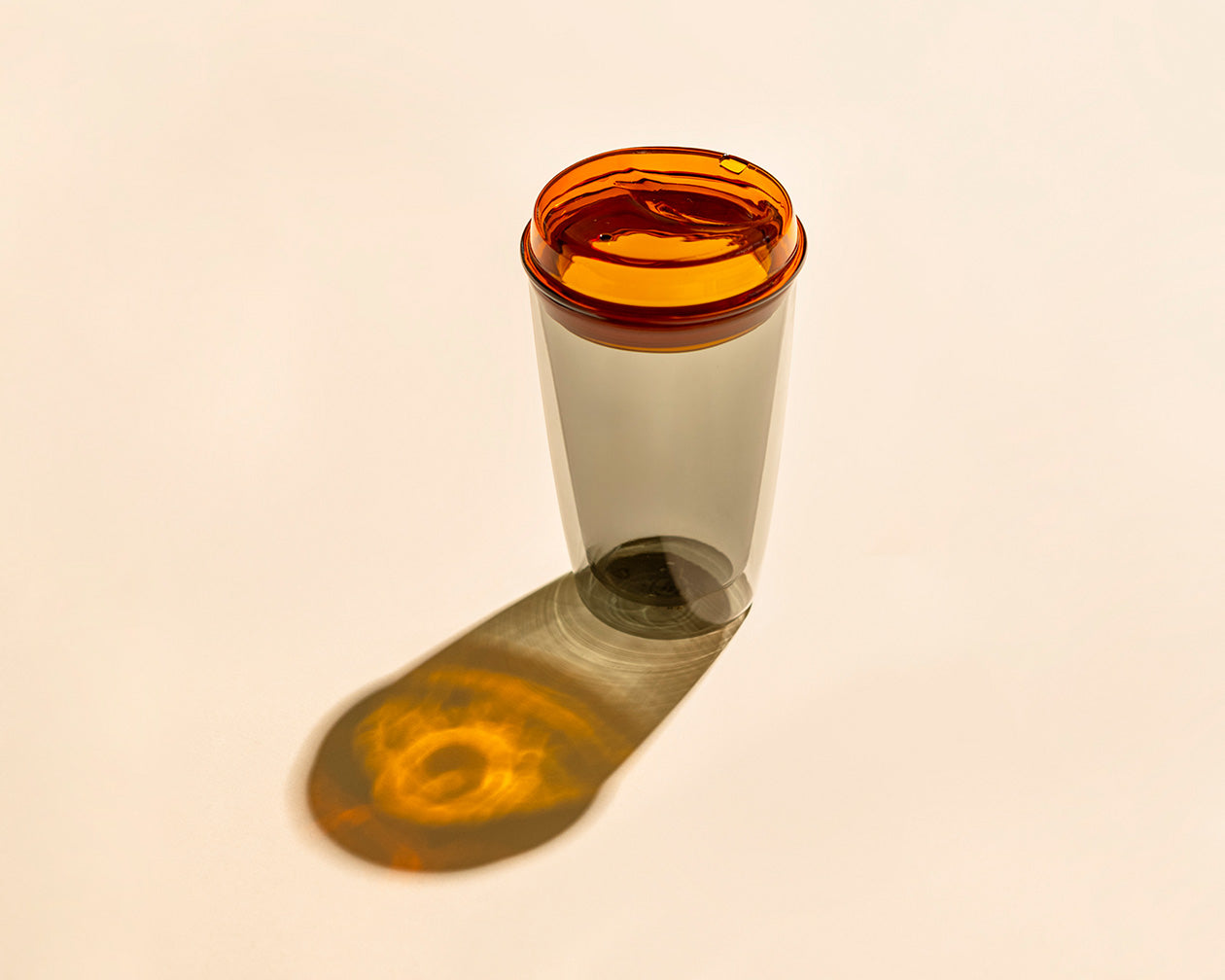 A translucent amber-tinted plastic cup with a matching amber lid, standing on a warm off-white background and casting a long shadow.