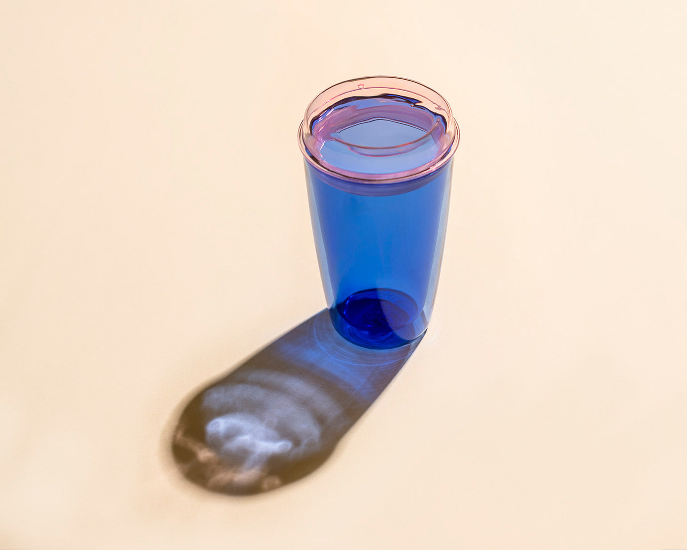A translucent blue plastic tumbler with a pink rim, standing on a beige surface and casting a long shadow.