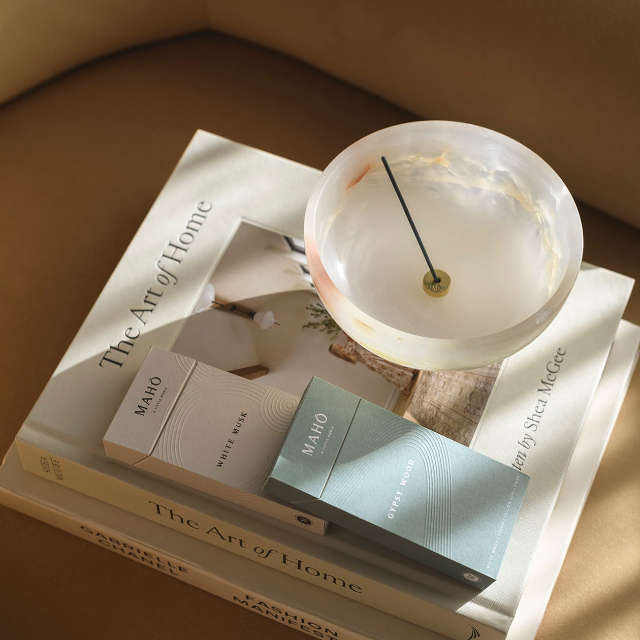 A styled interior scene showing a stack of books on a beige upholstered chair, with a round translucent white vessel and a small rectangular box placed on top. The books include visible titles such as 'The Art of Home' and 'The Art of Living'; the box appears branded 'MAMO'.