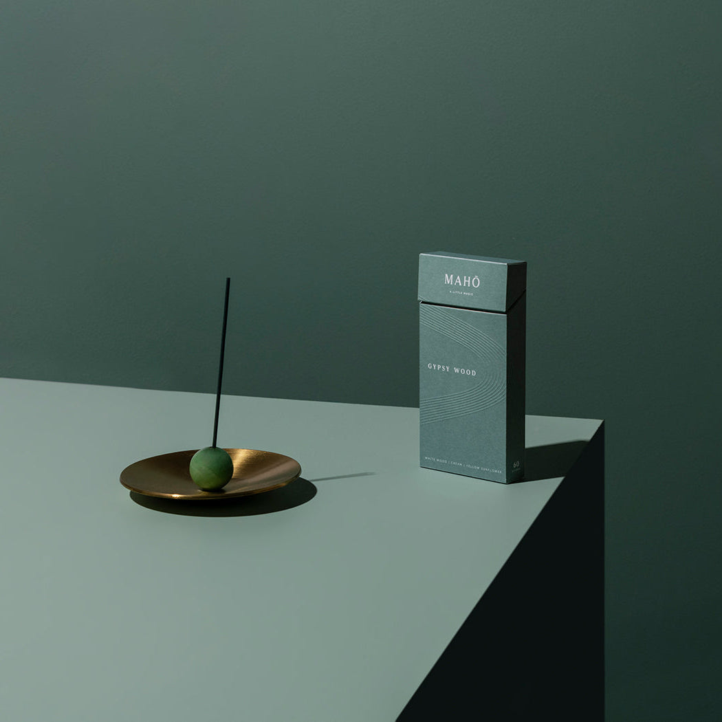 A slim rectangular MAHO incense box in a muted blue-gray color stands on a white platform against a dark green wall. To the left, a small gold dish holds a green sphere with a thin black incense stick.