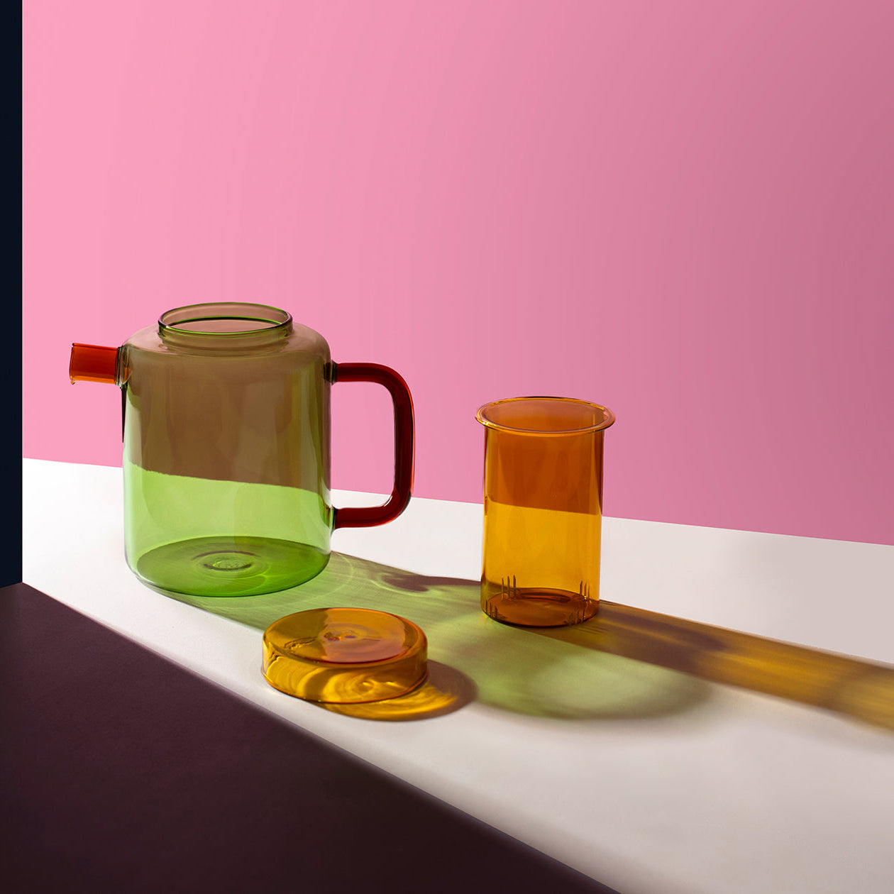 A translucent green teapot with a red handle and spout, shown alongside a matching amber glass cup and lid on a white surface against a pink background.