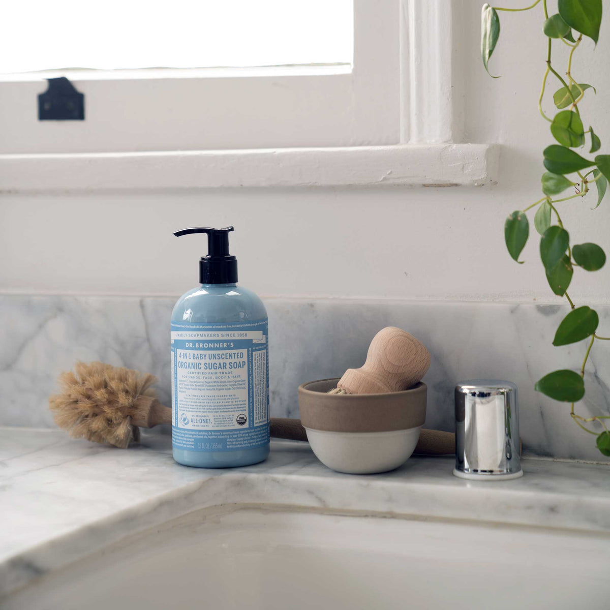 Dr. Bronner's Baby Unscented Organic Sugar Soap pump bottle on a marble basin with a wooden brush and trailing plant