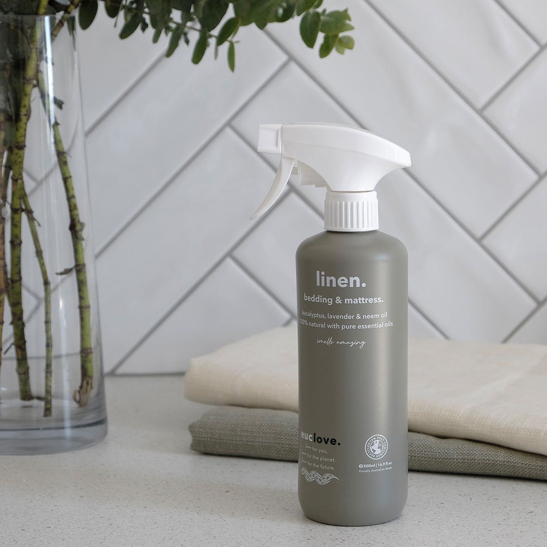 Euclove linen, bedding and mattress spray bottle beside folded linen sheets and a vase of foliage